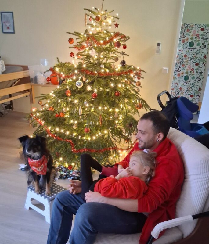 Ein Pfleger sitzt mit einem kleinem Mädchen und einem Therapiehund vor einem leuchtenden Weihnachtsbaum.